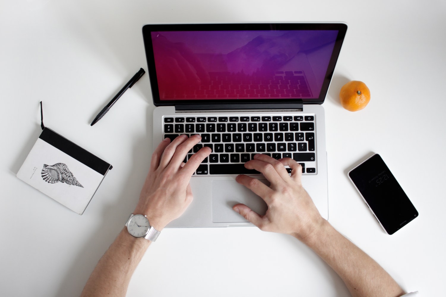 An image of a person using an apple mac laptop with an apple, iphone, pen and nodepad surrounding
